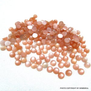 May include: A large pile of round, peach-colored sunstone cabochons. The stones have a shimmery, iridescent finish.