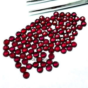 Könnte beinhalten: Eine Ansammlung facettierter, tiefroter Edelsteine, die auf einer weißen Oberfläche verstreut sind. Die Edelsteine sind rund mit mehreren Facetten, die das Licht reflektieren. Eine silberne Pinzette befindet sich oben. Der Text "GEMSDEL" ist sichtbar.