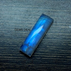 Puede incluir: Una gema rectangular pulida con un tono azul vibrante, con reflejos internos de luz. La piedra está sobre una superficie oscura y texturizada. El texto "GEMS DEAL" es visible.