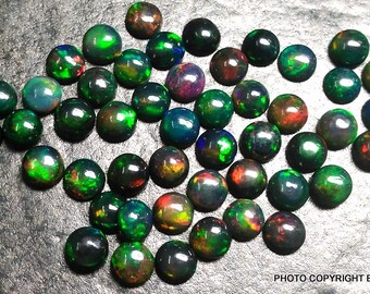 May include: A collection of round, black opal cabochons with green, red, and blue iridescent flashes. The opals are arranged in a random pattern on a gray surface.