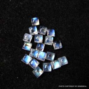K&ouml;nnte beinhalten: Eine Gruppe von 17 regenbogenfarbenen blauen Mondstein-Cabochons, die jeweils in quadratischer Form geschnitten sind. Die Steine liegen lose auf einem schwarzen Hintergrund.