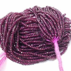 May include: A strand of deep red faceted gemstone beads. The beads are small and uniform in size, and they are strung on a thin thread. The beads are tied together with a pink thread at the end.