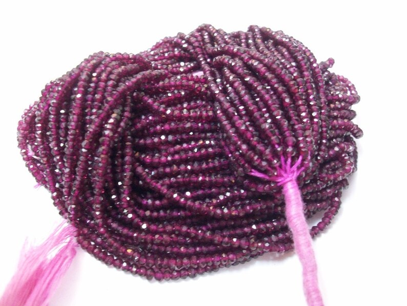 May include: A strand of deep red faceted gemstone beads. The beads are small and uniform in size, and they are strung on a thin thread. The beads are tied together with a pink thread at the end.