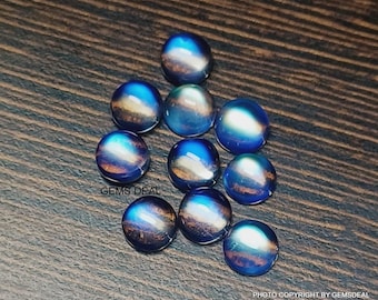 Op de afbeelding: Een verzameling iriserende, ronde edelstenen met een gepolijste afwerking. De edelstenen vertonen een mix van blauwe, gouden en paarse tinten, die het licht reflecteren. De tekst "GEMS DEAL" is zichtbaar. De edelstenen liggen op een donker houten oppervlak.