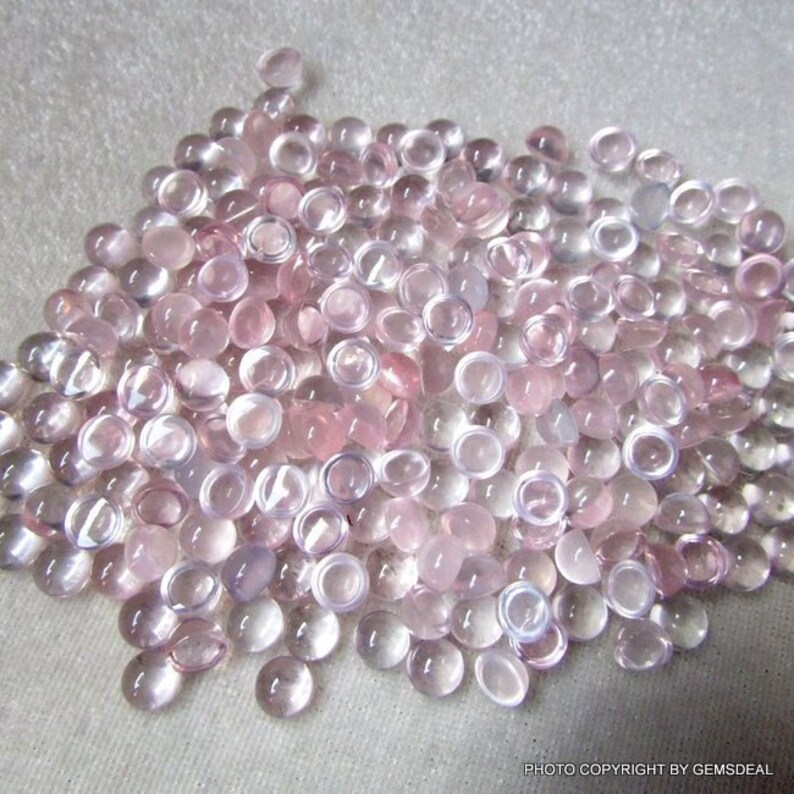 May include: A large pile of pale pink and clear round cabochon gemstones. The gemstones are smooth and polished, and they have a slightly translucent appearance.