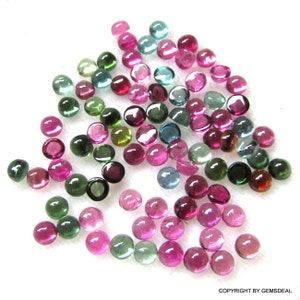 May include: A collection of round, faceted gemstones in various shades of green, pink, and blue. The gemstones are scattered on a white surface.