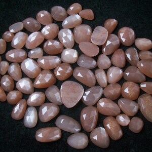 May include: A collection of approximately 40 sunstone gemstones, each with a unique, faceted shape. The stones are a pale peach color with a subtle shimmer.