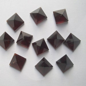 May include: Ten dark red square pyramid-shaped gemstones. The gemstones are arranged in a scattered pattern on a white surface.