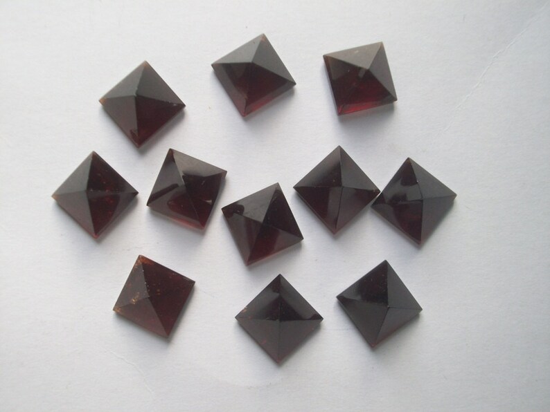 May include: Ten dark red square pyramid-shaped gemstones. The gemstones are arranged in a scattered pattern on a white surface.