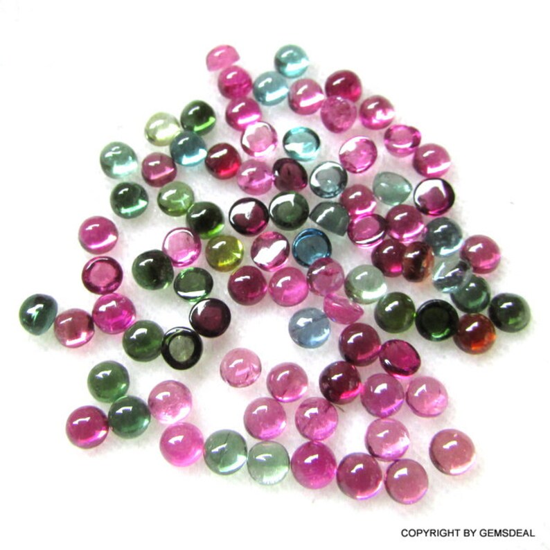 May include: A collection of round, faceted gemstones in various shades of green, pink, and blue. The gemstones are scattered on a white surface.