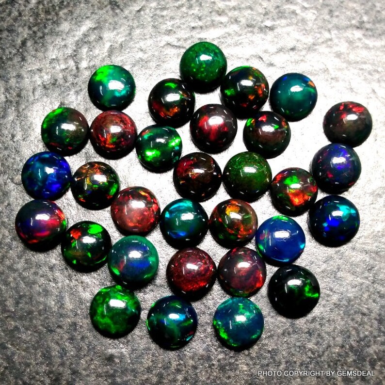 May include: A collection of 25 round, black opal cabochons with iridescent green, blue, and red flashes. The opals are arranged in a random pattern on a gray surface.