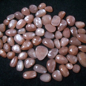 May include: A collection of approximately 40 sunstone cabochons in various sizes and shapes. The stones are a light peach color with a shimmery, iridescent finish.