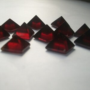 May include: A group of ten red, faceted, pyramid-shaped gemstones. The gemstones are arranged in a random pattern on a white surface.