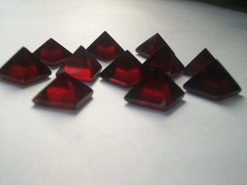 May include: A group of ten red, faceted, pyramid-shaped gemstones. The gemstones are arranged in a random pattern on a white surface.