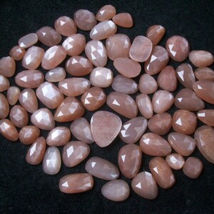 May include: A collection of approximately 40 sunstone cabochons in various sizes and shapes. The stones are a light peach color with a shimmery, iridescent finish.