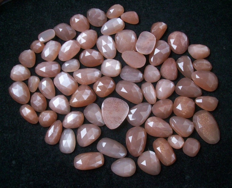 May include: A collection of approximately 40 sunstone cabochons in various sizes and shapes. The stones are a light peach color with a shimmery, iridescent finish.