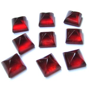 May include: Nine square-shaped, faceted beads in a deep red color. The beads have a pyramid-like design on each side, with a lighter red center. They are arranged on a white background.