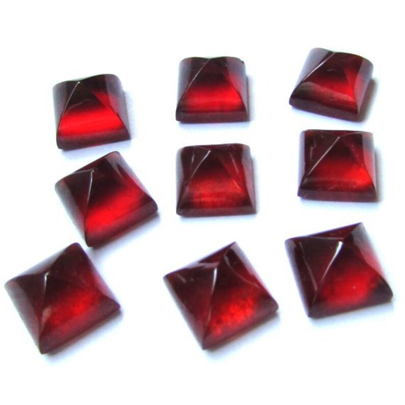May include: Nine square-shaped, faceted beads in a deep red color. The beads have a pyramid-like design on each side, with a lighter red center. They are arranged on a white background.