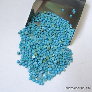 May include: A collection of small, round, turquoise-colored gemstones spilling from a metal scoop onto a white surface. The gemstones have a smooth, polished appearance and vary slightly in shade, with some showing hints of green.