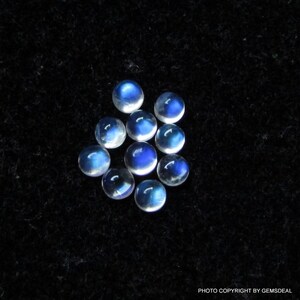 May include: A group of ten round, iridescent moonstone cabochons, each with a blue sheen. The stones are arranged in a loose cluster on a black background.