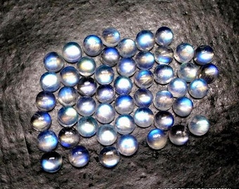 Puede incluir: Una colección de cabochones de piedra lunar azul iridiscente, cada uno con un brillo y un brillo únicos. Las piedras están dispuestas en un montón suelto sobre un fondo oscuro.