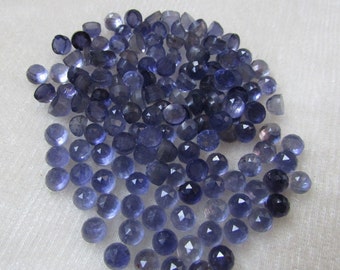 May include: A pile of approximately 70 small, faceted, round, blue gemstones. The gemstones are arranged in a loose pile on a white surface.