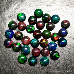 May include: A collection of 25 round, black opal gemstones with iridescent colors, including green, red, blue, and purple. The opals are arranged in a random pattern on a gray surface.