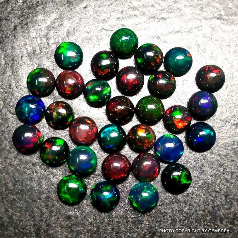 May include: A collection of 25 round, black opal gemstones with iridescent colors, including green, red, blue, and purple. The opals are arranged in a random pattern on a gray surface.