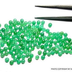 May include: A large group of round, green emerald gemstones. The gemstones are scattered on a white surface.