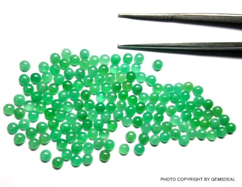 May include: A large group of round, green emerald gemstones. The gemstones are scattered on a white surface.