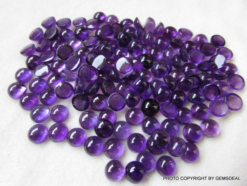 May include: A large pile of round, purple amethyst cabochons. The cabochons are smooth and polished, with a glossy finish.