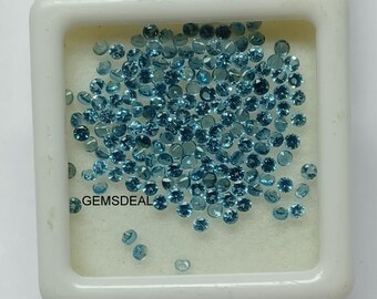 Op de afbeelding: Een verzameling kleine, ronde, gefacetteerde blauwe edelstenen in een vierkante witte opbergdoos. De tekst GEMSDEAL is in het zwart gedrukt op de witte schuimrubberen inzet. De stenen liggen verspreid over het oppervlak.
