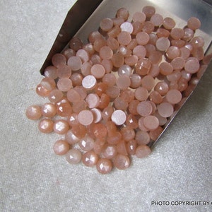 May include: A large pile of round, peach-colored sunstone cabochons in a metal tray. The stones have a shimmery, iridescent quality.