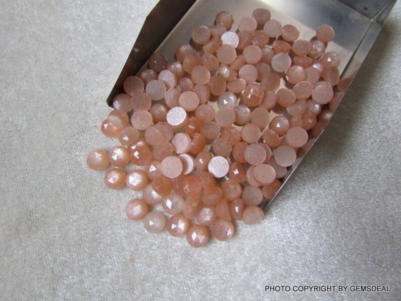 May include: A large pile of round, peach-colored sunstone cabochons in a metal tray. The stones have a shimmery, iridescent quality.