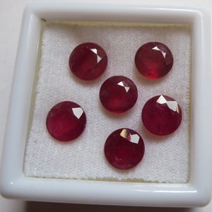 Puede incluir: Seis piedras preciosas redondas, facetadas, de color rubí rojo en una caja de presentación cuadrada blanca.