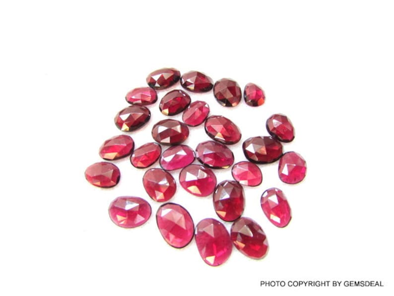 May include: A collection of 20 faceted, oval-shaped, red gemstones. The gemstones are scattered on a white surface.