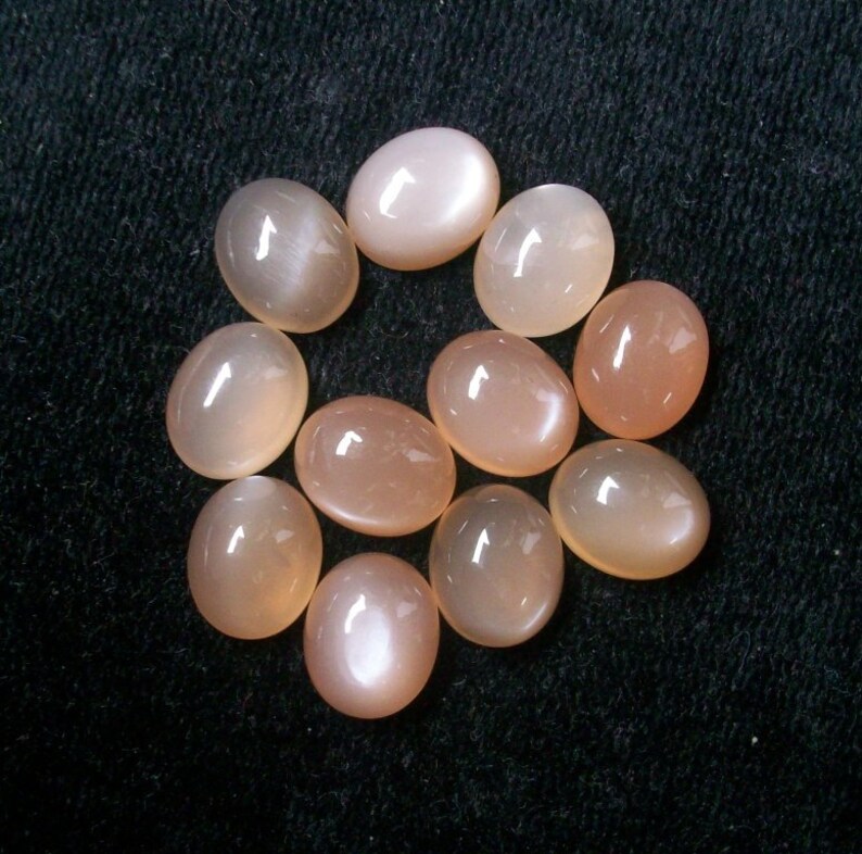 May include: A collection of ten oval-shaped moonstone cabochons in a pale peach color. The stones have a smooth, polished surface and a subtle shimmer.