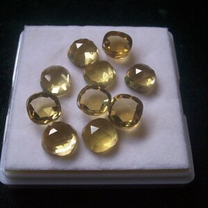 May include: A collection of ten cushion-cut, faceted, yellow gemstones displayed on a white background.