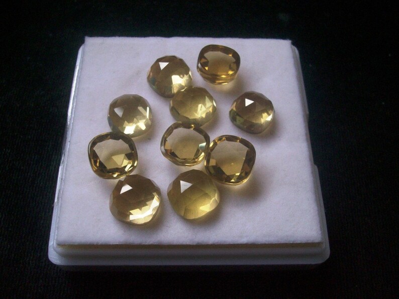 May include: A collection of ten cushion-cut, faceted, yellow gemstones displayed on a white background.