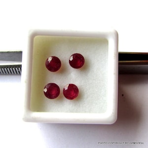 Könnte beinhalten: Vier runde, rote Rubin-Edelsteine in einer weißen quadratischen Box.