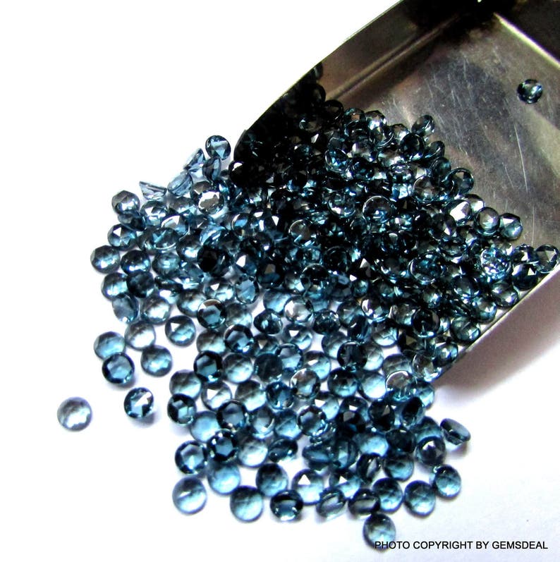 K&ouml;nnte beinhalten: Eine Schaufel mit losen blauen Topas-Edelsteinen, im Hintergrund eine silberne Schaufel. Die Edelsteine sind rund und facettiert.
