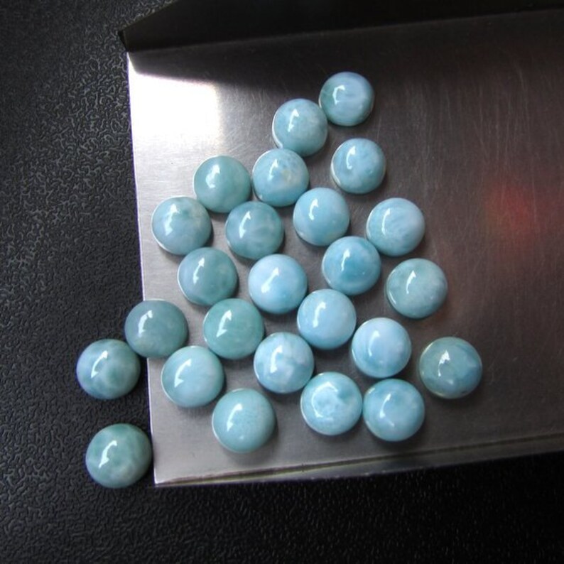 May include: A collection of round, light blue cabochon gemstones with a cloudy, marbled pattern. The stones are arranged in a triangular shape on a silver tray.