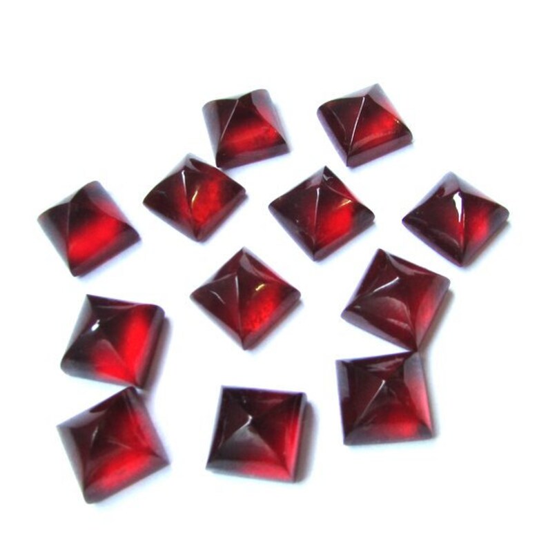 May include: A collection of square, faceted, dark red gemstones. Each gem has a pyramidal shape with a glossy finish, reflecting light. The gems are scattered against a white background, showcasing their rich color and intricate cuts. Ideal for jewelry making or crafts.