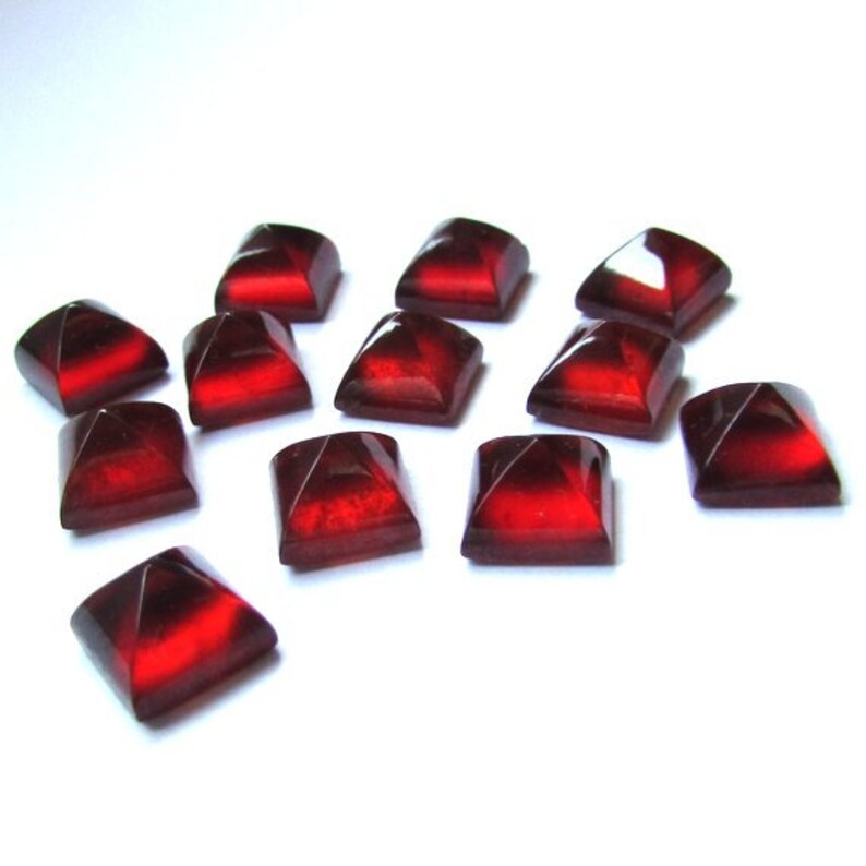 May include: A collection of square-shaped, faceted beads in a deep red color. The beads have a glossy finish and are scattered on a white surface. The facets create a play of light and shadow, enhancing their jewel-like appearance.