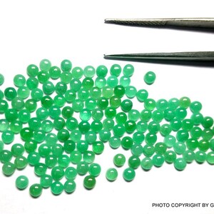 May include: A large group of round, green emerald gemstones. The gemstones are scattered on a white surface.