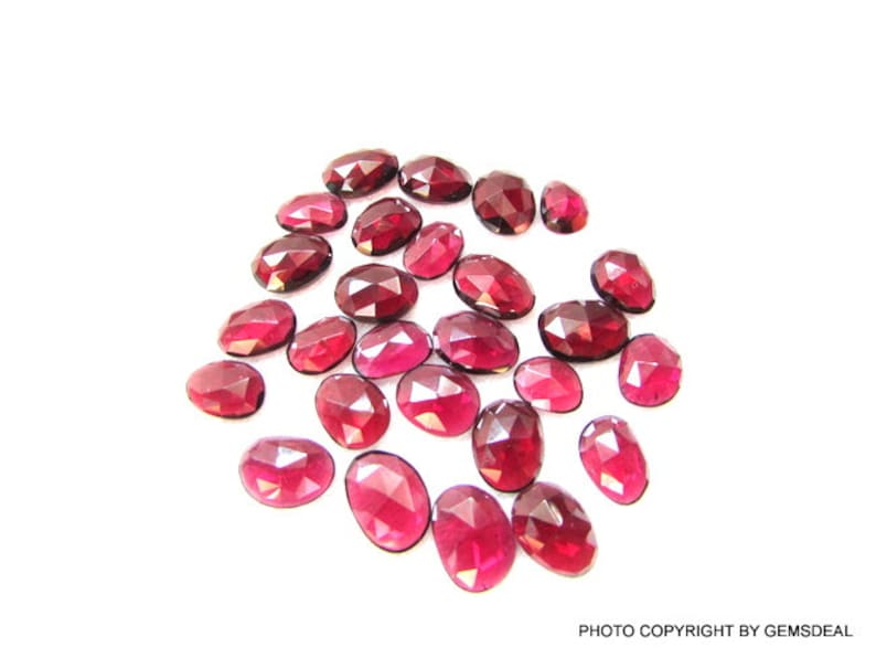 May include: A collection of 20 oval-shaped, faceted, red gemstones. The gemstones are loose and scattered on a white surface.