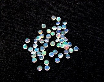 Pode incluir: Uma coleção de gemas de opala branca iridescente, cada uma com um corte redondo em cabochão. As opalas estão espalhadas sobre uma superfície preta.