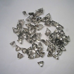 May include: A collection of small, triangular, clear quartz crystals with black inclusions. The crystals are scattered on a white surface.