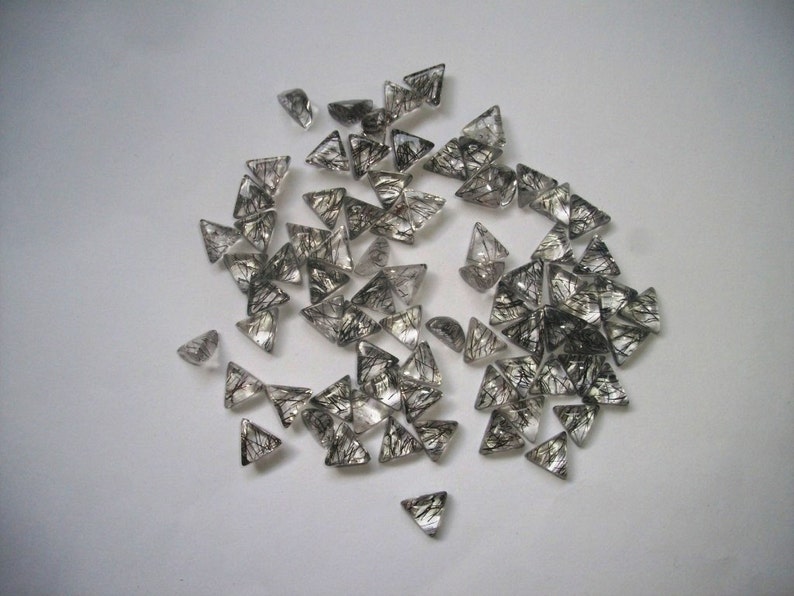 May include: A collection of small, triangular, clear quartz crystals with black inclusions. The crystals are scattered on a white surface.