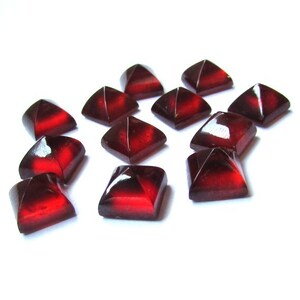 May include: A collection of faceted, square-shaped gemstones in a deep red color. The gems have a glossy finish and are arranged on a white surface, with light reflecting off their surfaces. These could be used for jewelry making or other crafts.